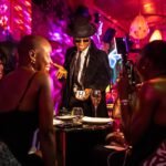 Mágico com chapéu e óculos escuros executa truque de cartas para grupo de mulheres sentadas em mesa de bar com iluminação colorida.