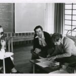 Quatro adultos sentados em sala discutem documentos sobre mesa baixa. Mulher à esquerda usa óculos escuros e segura papel. Três homens à direita observam e escrevem em papéis. Quadro-negro com anotações ao fundo e janela com cortinas à direita.