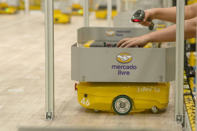 A imagem mostra um robô de armazenamento do Mercado Livre em um armazém. O robô é de cor amarela e possui um compartimento cinza com o logotipo do Mercado Livre. Uma mão humana está segurando um scanner, apontando para o robô. Ao fundo, outros robôs semelhantes estão visíveis em um ambiente de armazém com piso de madeira.