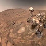 Rover com rodas e instrumentos científicos em solo marciano acidentado, com colinas e céu alaranjado ao fundo.