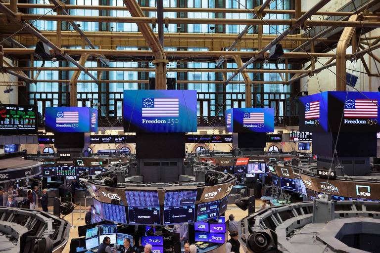 Área de negociação da Bolsa de Valores de Nova York com múltiplos monitores exibindo a bandeira americana e a mensagem 'Freedom 250'. Estrutura de madeira e janelas altas ao fundo.