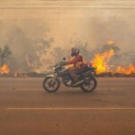 Motociclista com capacete e colete vermelho trafega em estrada asfaltada enquanto fogo consome vegetação seca ao fundo. Fumaça densa cobre o céu e árvores parcialmente queimadas são visíveis.