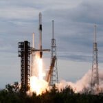 Foguete Falcon 9 da SpaceX decola da plataforma de lançamento com chamas e fumaça intensa na base. Estruturas metálicas altas cercam a plataforma sob céu nublado.