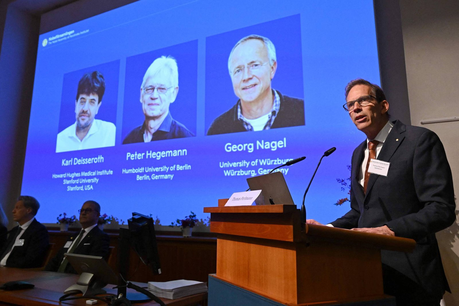 Homem fala em púlpito com microfone em auditório. Ao fundo, tela exibe fotos e nomes de três pesquisadores: Karl Deisseroth, Peter Hegemann e Georg Nagel, com suas instituições e países.
