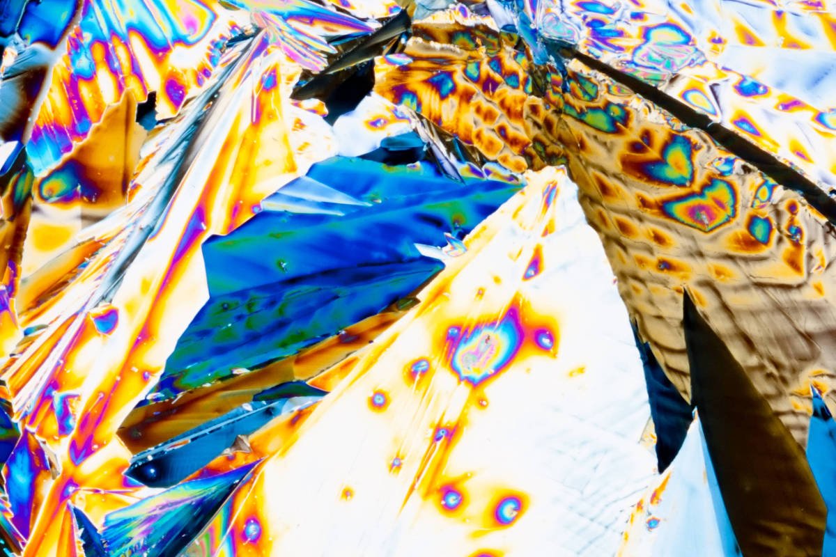 Imagem macro mostra cristais com padrões iridescentes em tons de azul, laranja e amarelo, exibindo texturas facetadas e detalhes vibrantes.