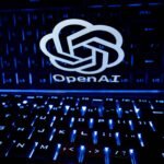 Logo branco da OpenAI refletido na superfície brilhante acima de teclado retroiluminado em azul. Teclas com letras e símbolos visíveis em ambiente escuro.