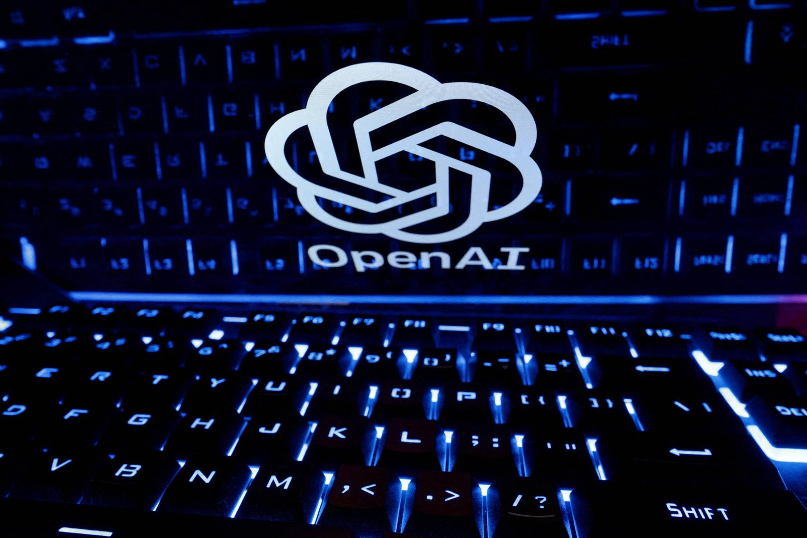 Logo branco da OpenAI refletido na superfície brilhante acima de teclado retroiluminado em azul. Teclas com letras e símbolos visíveis em ambiente escuro.
