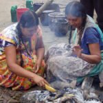 Duas mulheres indígenas sentadas no chão preparam alimentos em uma fogueira ao ar livre. Elas usam vestidos coloridos e têm cabelos trançados. Ao fundo, utensílios e recipientes estão dispostos sobre o solo.