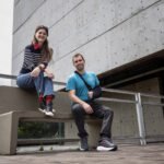 Uma mulher com blusa listrada preta e branca, calça jeans e tênis pretos está sentada no encosto de um banco de concreto. Ao lado dela, um homem com camiseta azul, calça preta e tênis cinza está sentado no assento do banco, com o braço direito imobilizado por tipoia preta. Ambos estão em área externa com piso de pedra, próximo a um prédio de concreto com janelas escuras e guarda-corpo metálico.