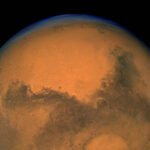 Imagem do planeta Marte mostrando sua superfície avermelhada com áreas escuras e uma calota polar branca na parte inferior.